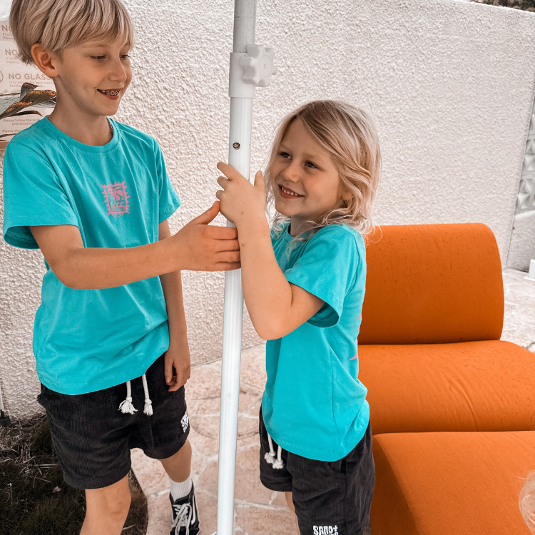 Two children in matching turquoise shirts standing next to an orange chair against a textured white wall.