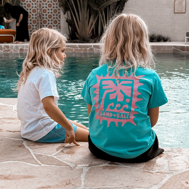 Two children sitting by a pool, one wearing a white shirt and the other a teal shirt with a logo.