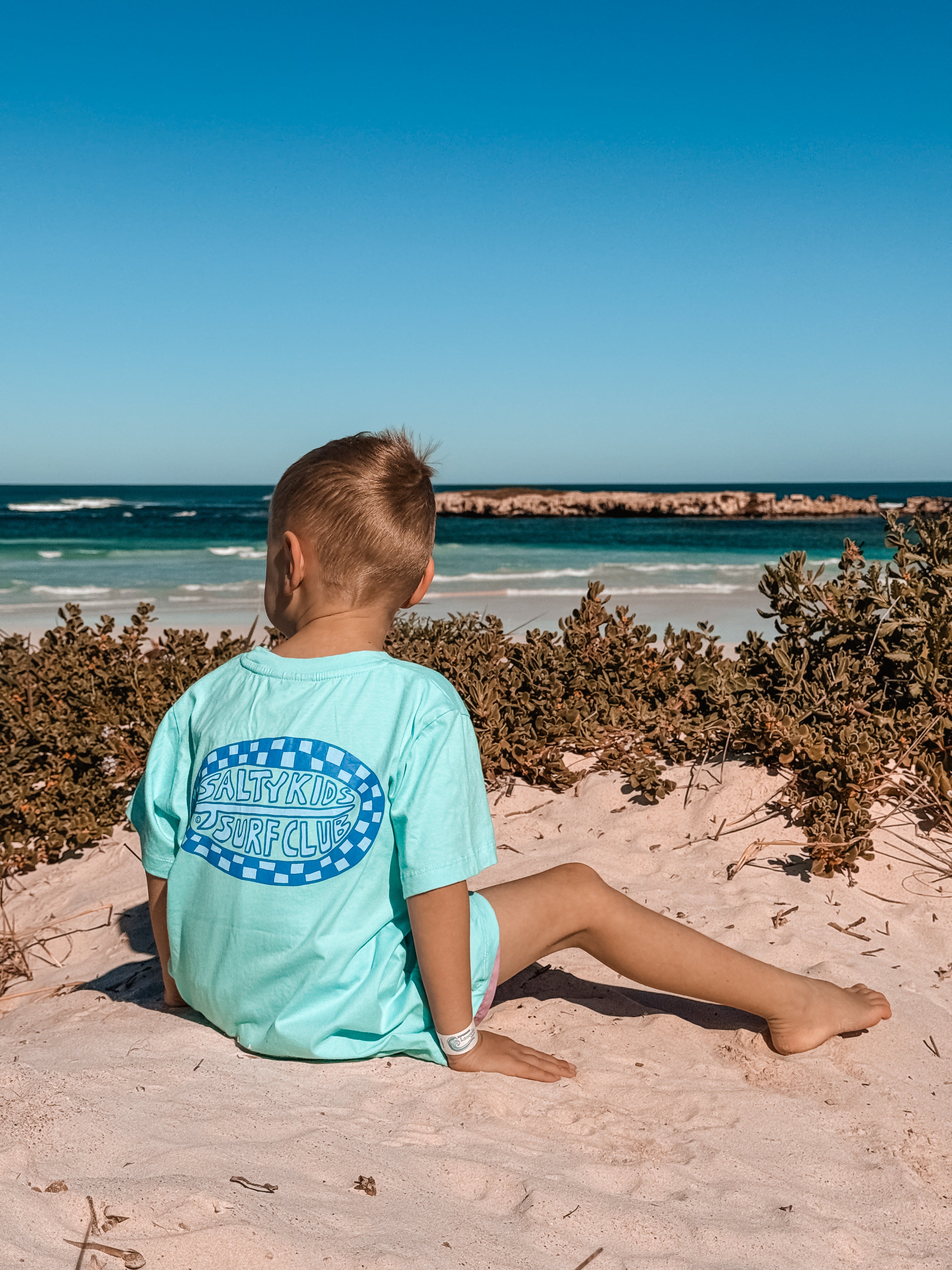 A child sitting on a sandy beach facing the ocean, wearing a light aqua t-shirt with a blue and white 'salty kids Surf Club' graphic on the back.