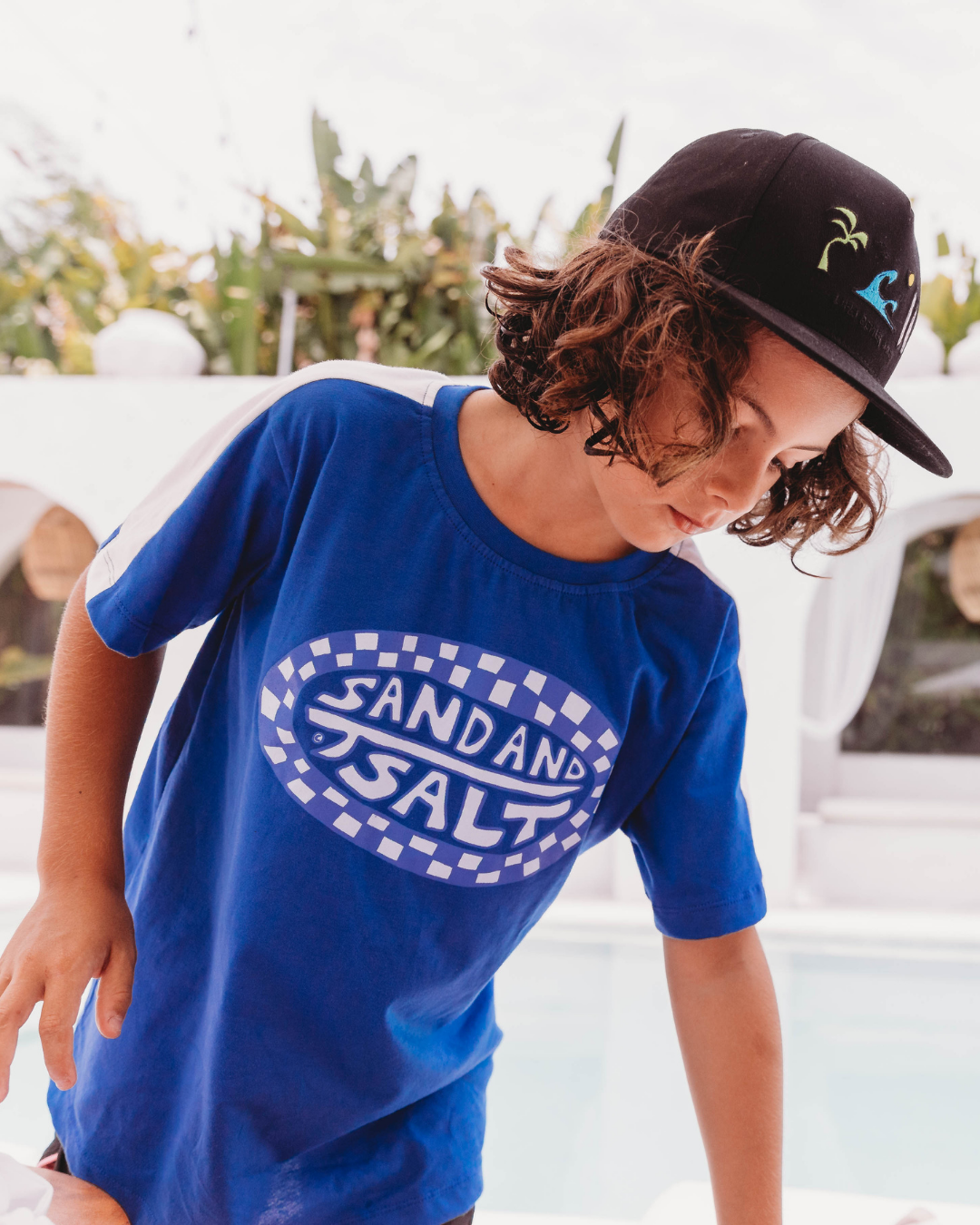 Boy leaning forward at poolside wearing bright blue Sand + Salt checker badge tee and black snapback, white arched wall background