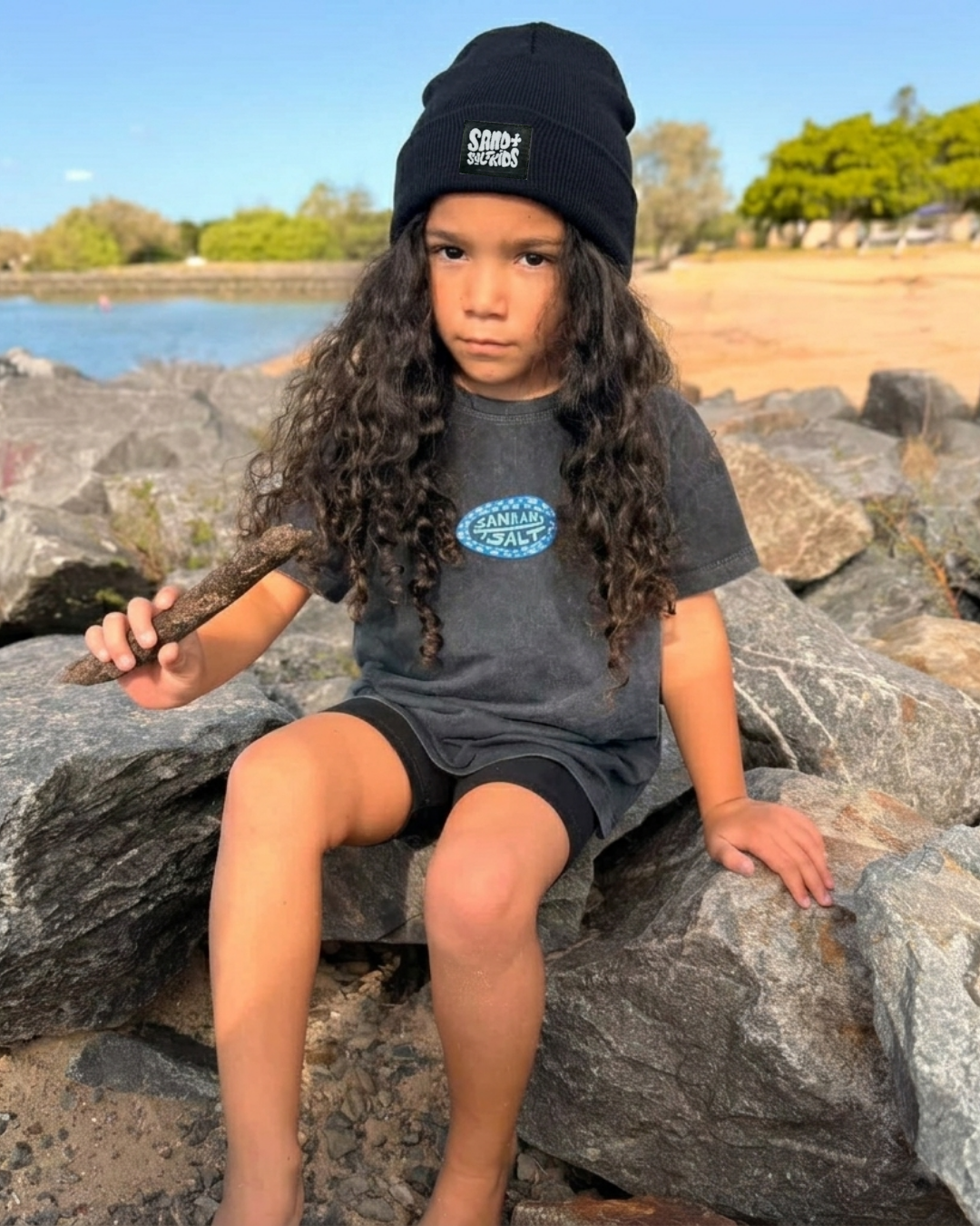 Child sitting on rocks by a lake, wearing a black beanie and gray shirt.