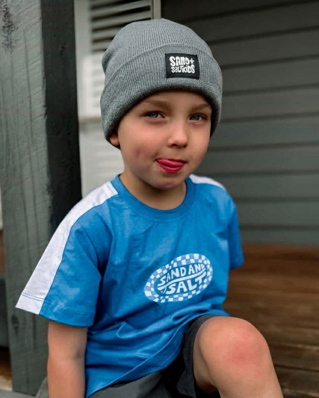 Child wearing a blue shirt and gray beanie with visible branding, sitting on a wooden step.