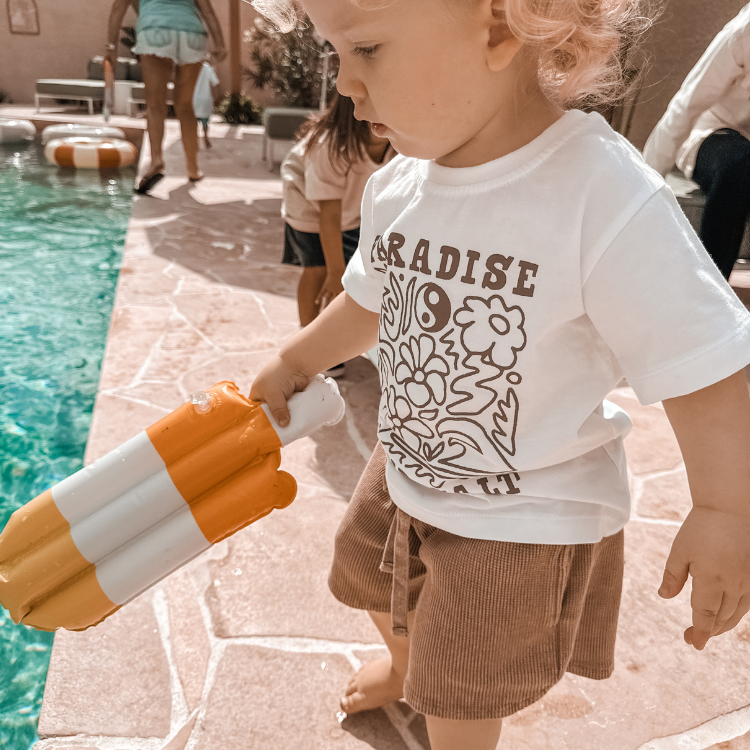 Child holding a colorful inflatable toy by a poolside wearing white tshirt paradise beach good vibes with waffle shorts