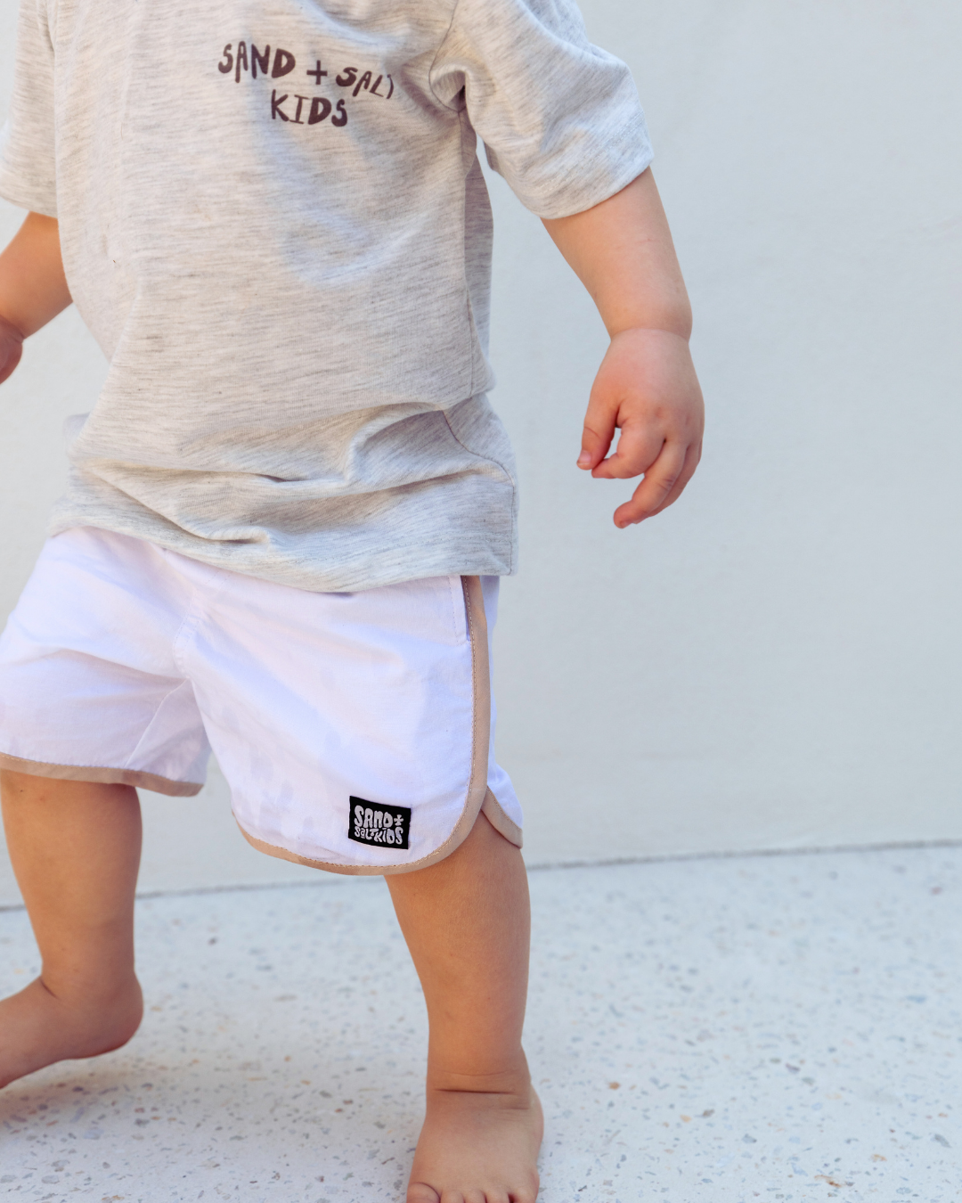 Child wearing Sand and Salt Kids white boardshorts, lightweight kids swim shorts designed for water play and everyday summer wear