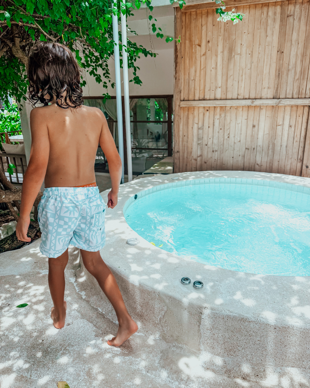 Boy walking towards round pool wearing Sand + Salt Kids Snapper Boardshorts, light blue and white checker graphic print, tropical Bali garden background