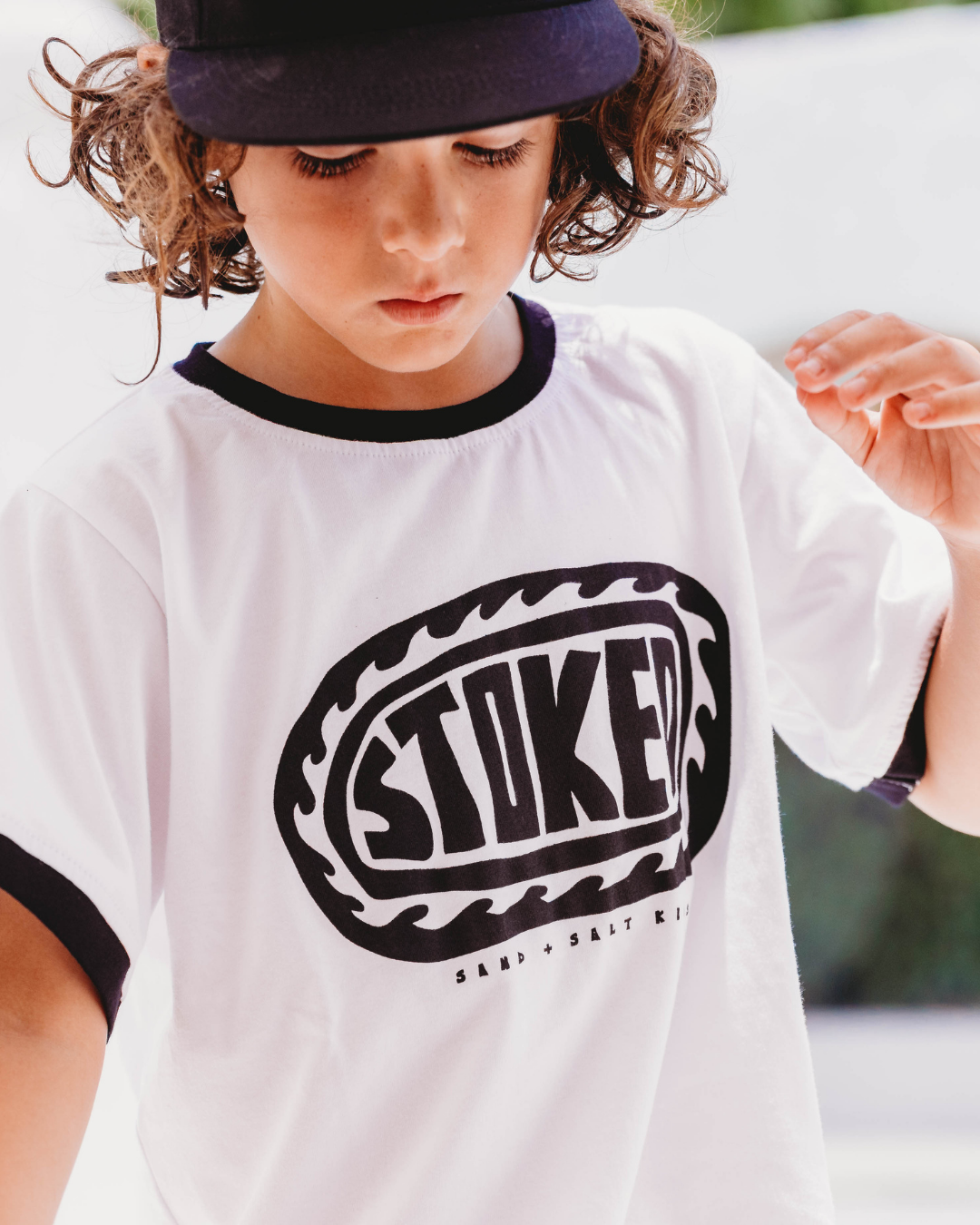 Boy sitting on white bench wearing white STOKED wave graphic ringer tee with black collar and cuffs, black snapback cap, white arched wall background