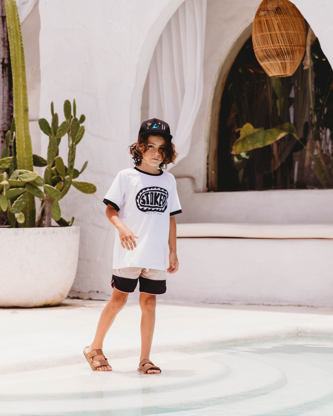 White kids tee with bold black STOKED wave graphic, folded on pool ledge with blurred pool and person in background, black rib collar visible, Sand + Salt Kids branding underneath graphic